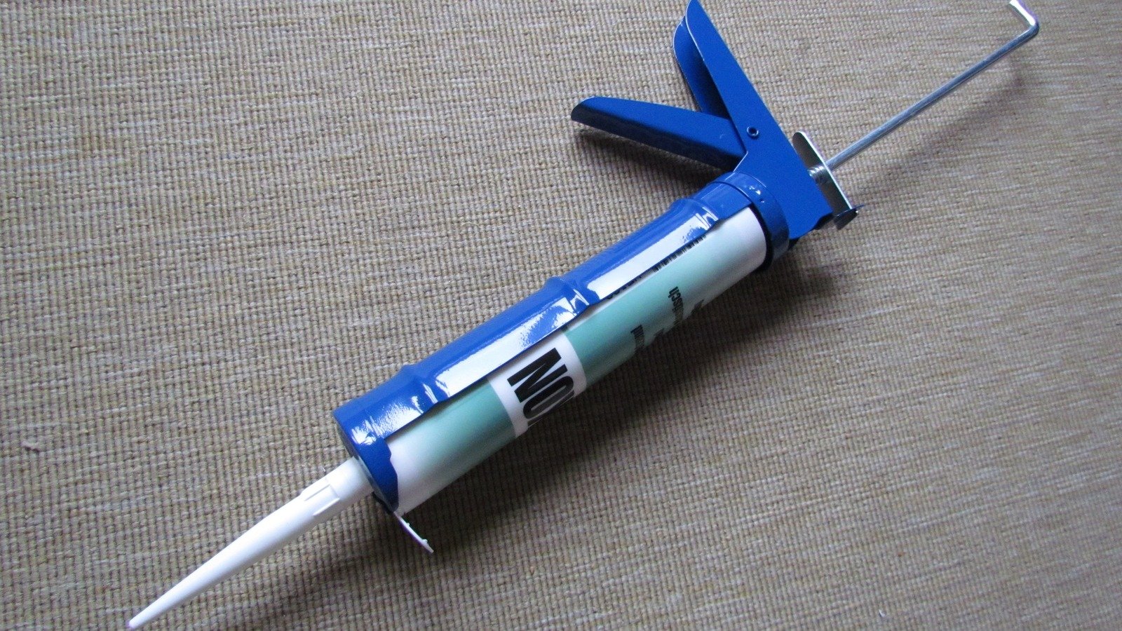 Blue caulking gun holding a sealant cartridge on a gray woven surface.