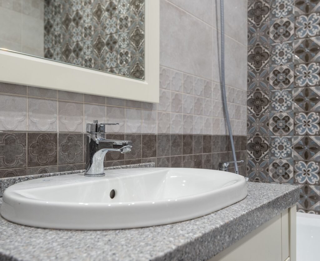 White bathroom sink beneath a chrome faucet, with patterned and gray tiled walls.