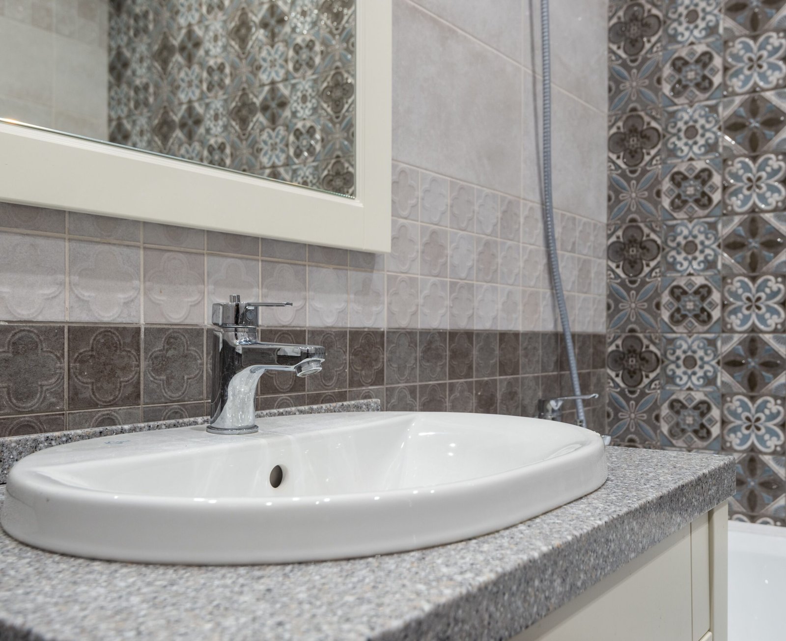 White bathroom sink beneath a chrome faucet, with patterned and gray tiled walls.