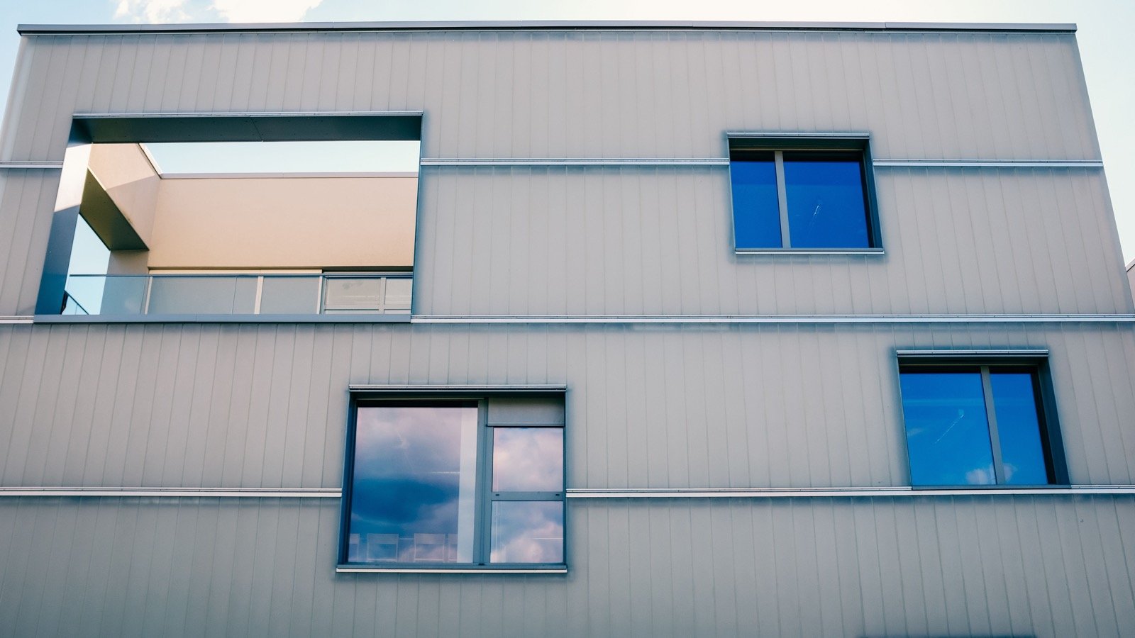 Modern residential facade with several windows and a recessed balcony beneath a pale blue sky.